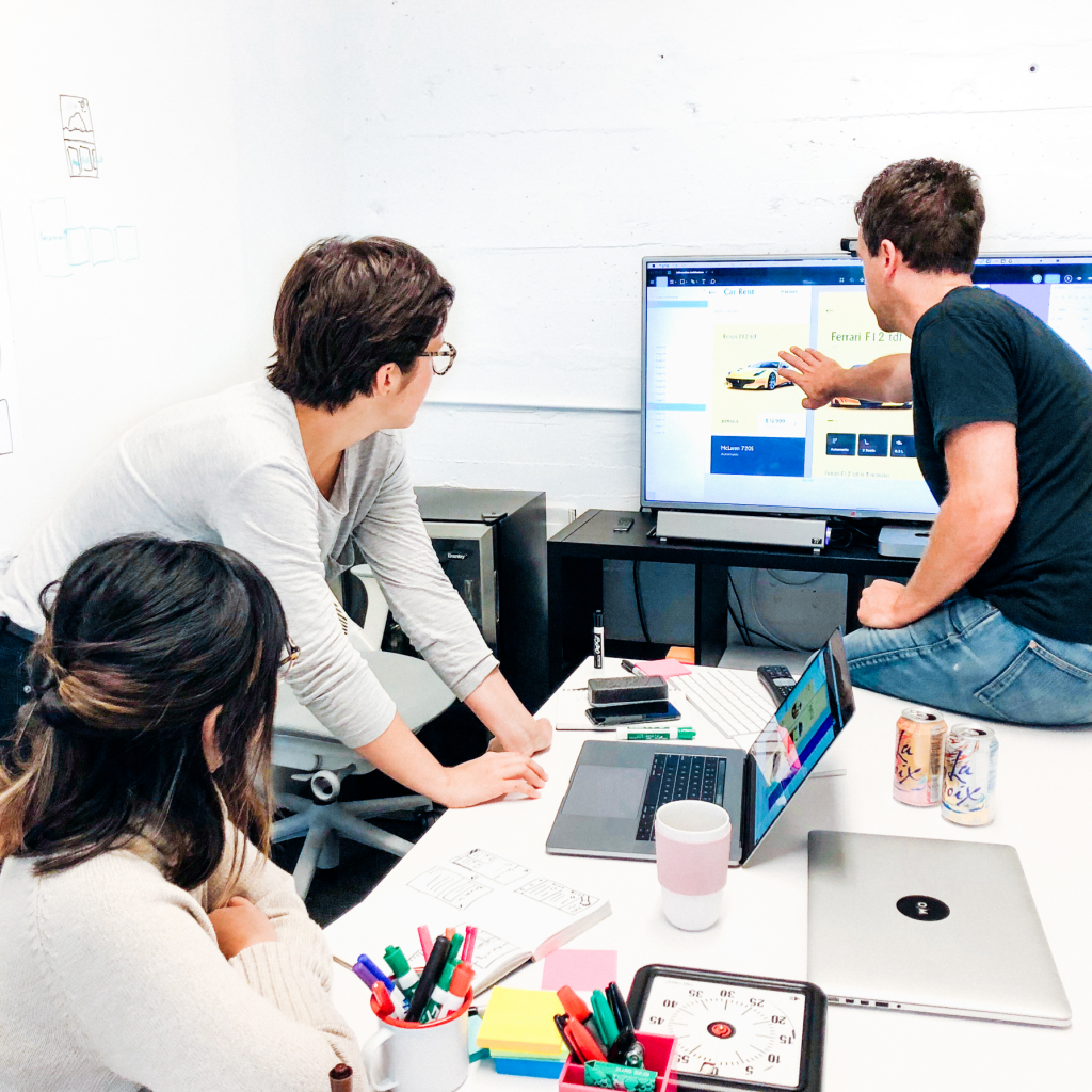 An image of a working session where three members of the design team, one man and two women, are reviewing recent work on a computer monitor. On the table are laptops, markers and post-it notes, and a meeting timer. 